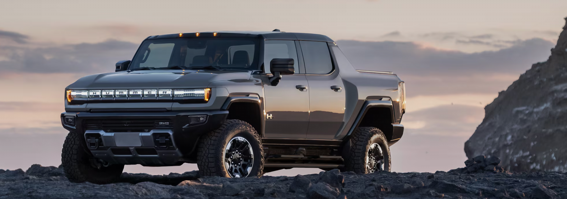 Front view of a 2026 GMC Hummer EV pickup truck parked on rocky terrain at sunset, showcasing its bold grille and off-road stance.