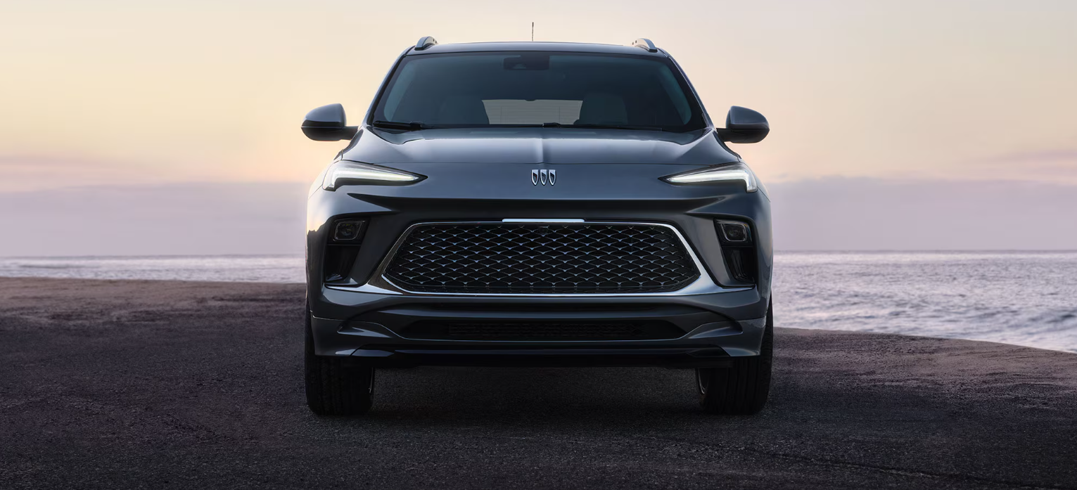 Front view of a 2026 Buick SUV parked by the ocean at sunset, showcasing its bold grille and modern LED headlights.